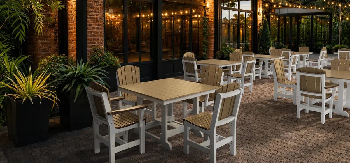 Weatherwood and white Wildridge poly dining tables and chairs set on a brick restaurant patio lit by string lights at dusk